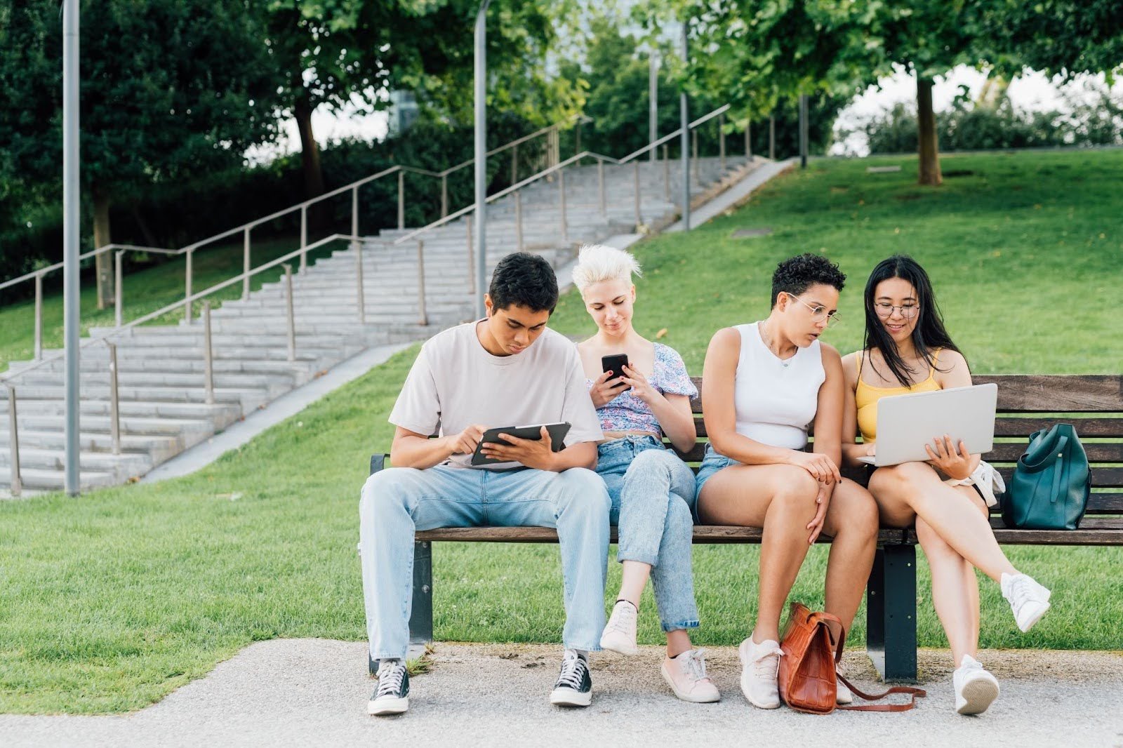 quatro amigos sentados no banco utilizando notebook e celular para aproveitar o programa de indicação.